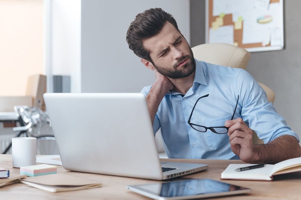 Man experiencing neck pain while sitting at a desk and working on a laptop